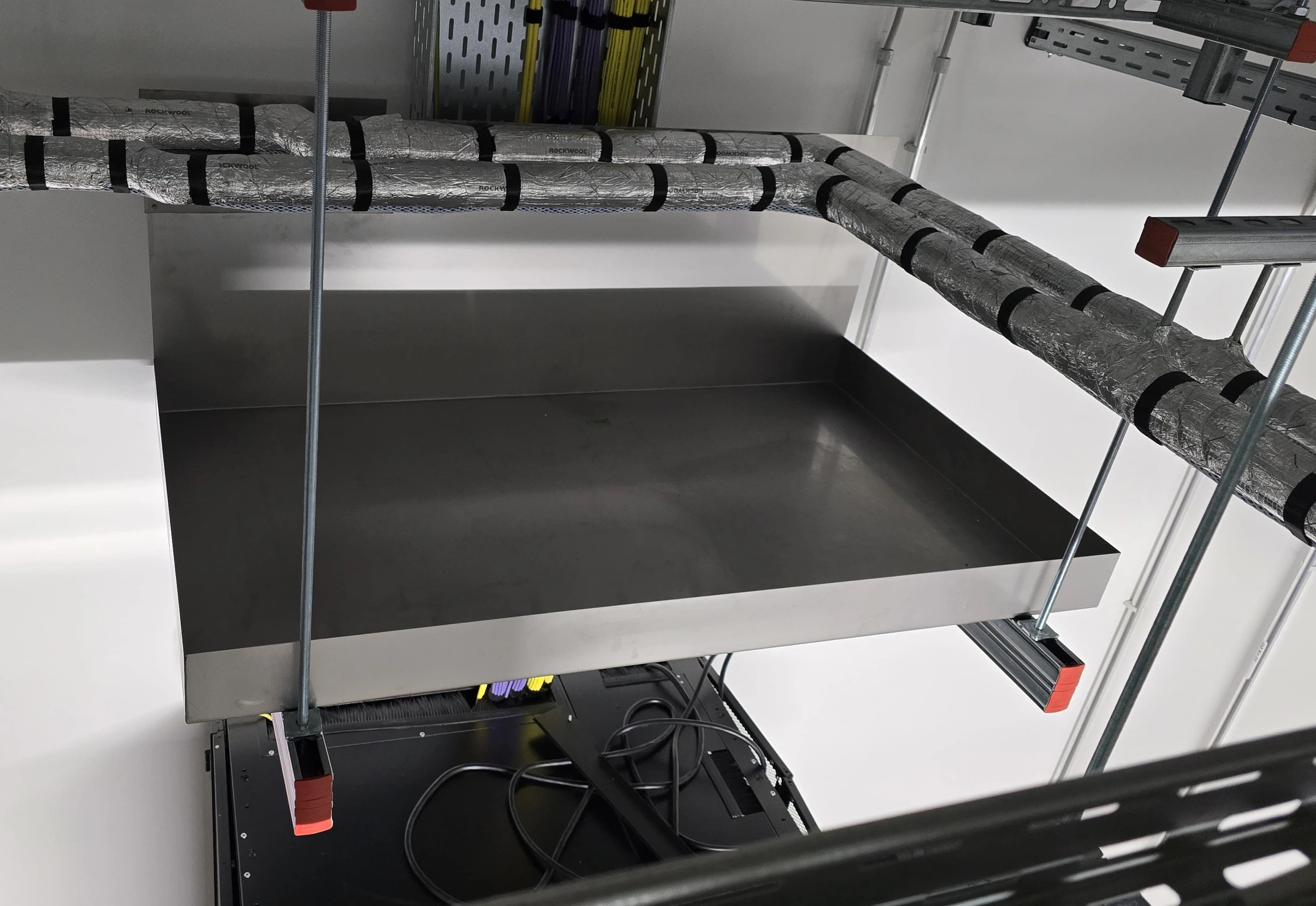 Sheet-metal drip tray beneath insulated pipework, supported by mounting rails and threaded rods. Sheet-metal drip tray beneath insulated pipework, supported by mounting rails and threaded rods.