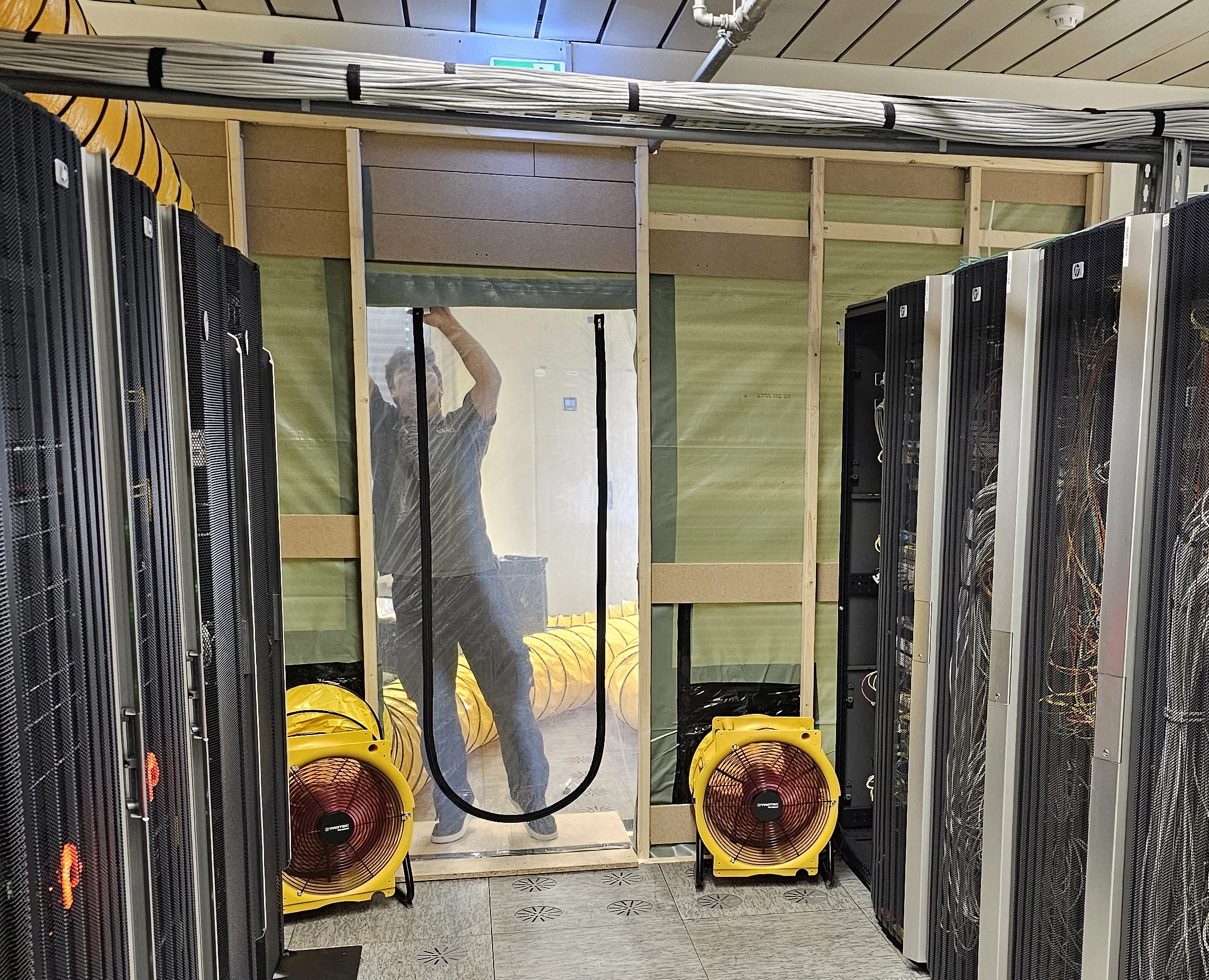 Team member at a temporary partition between rack rows with a plastic-sheet door, portable fans and air ducts.
