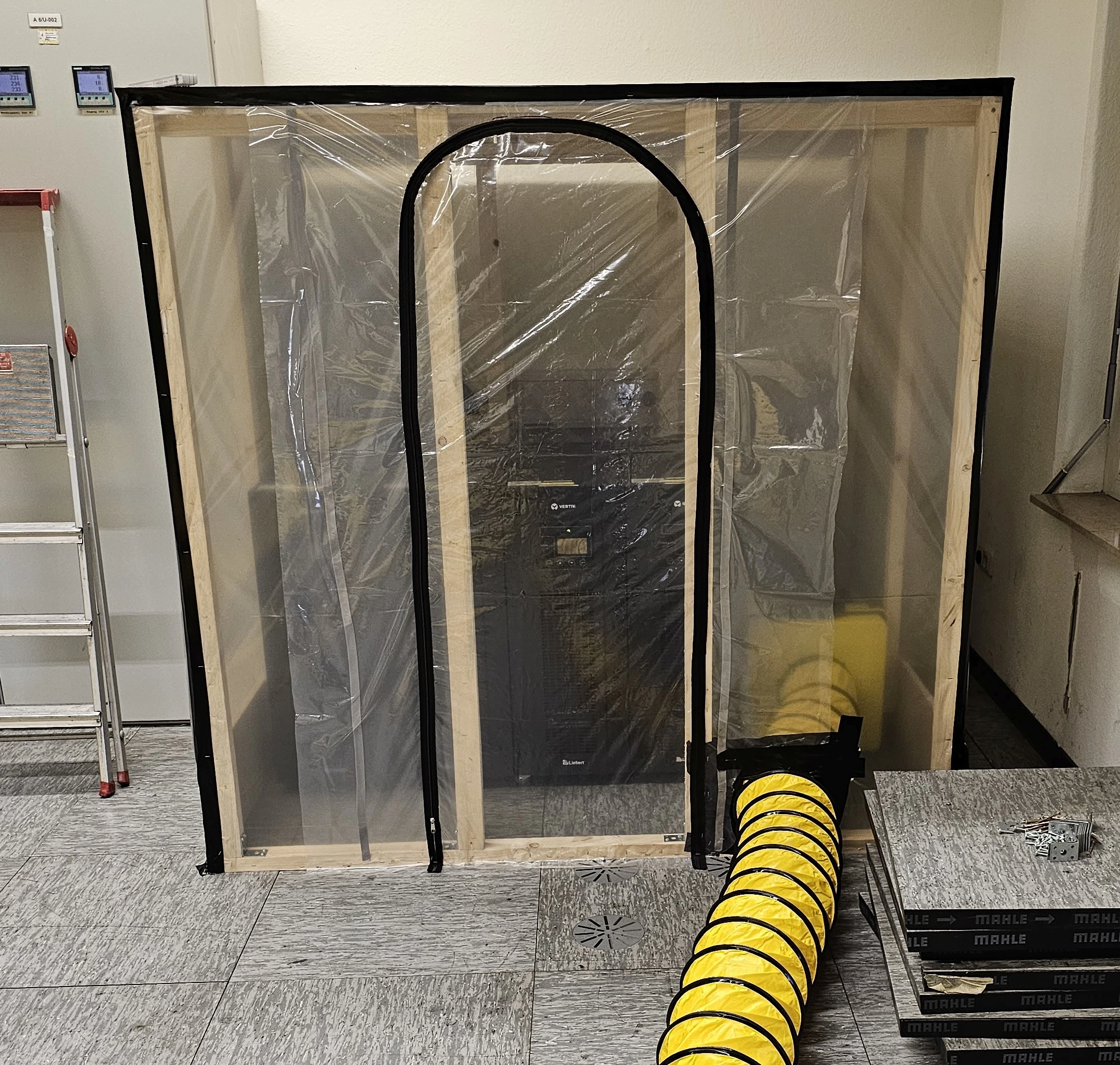 Timber-framed enclosure with transparent plastic sheeting, a zipped access door and a yellow air duct.