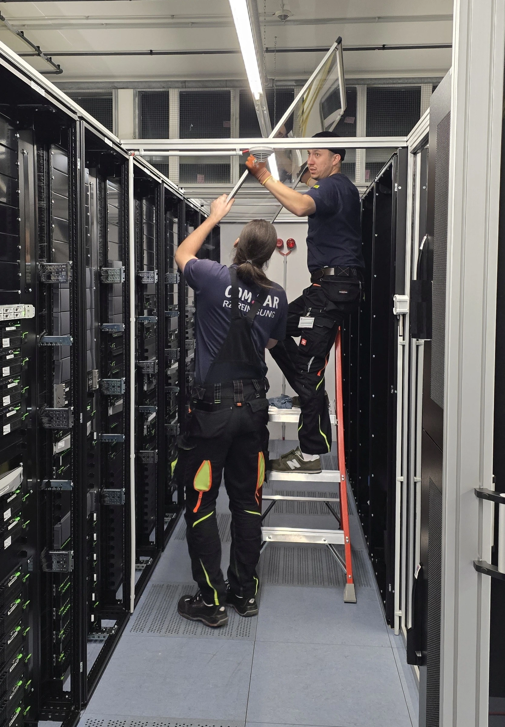Zwei Mitarbeitende reinigen die Einhausung über einem Gang zwischen Serverracks. Zwei Mitarbeitende reinigen die Einhausung über einem Gang zwischen Serverracks.
