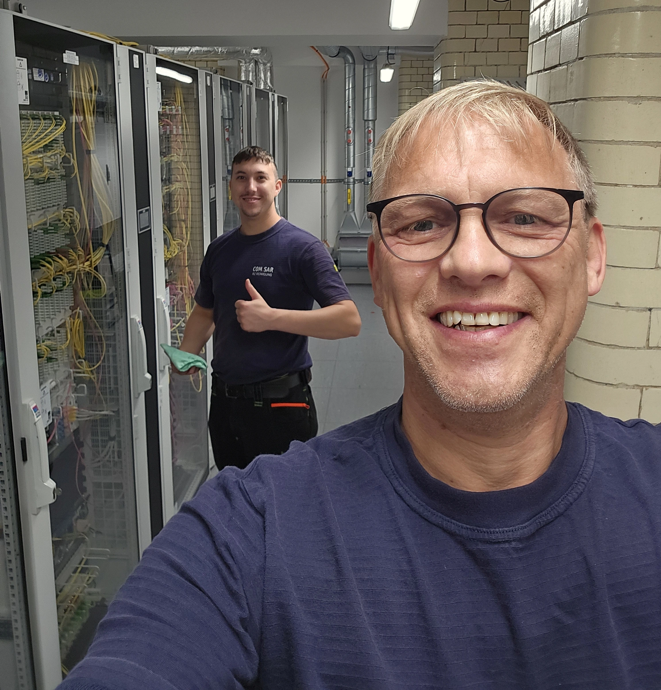 Zwei Mitarbeitende von COM.SAR IT Cleaning vor einer Rackreihe während eines Reinigungseinsatzes. Zwei Mitarbeitende von COM.SAR IT Cleaning vor einer Rackreihe während eines Reinigungseinsatzes.